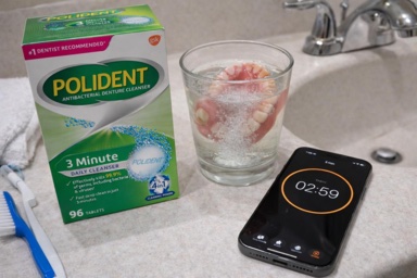 A box of Polident next to dentures soaking in a glass, with a smartphone timer counting down